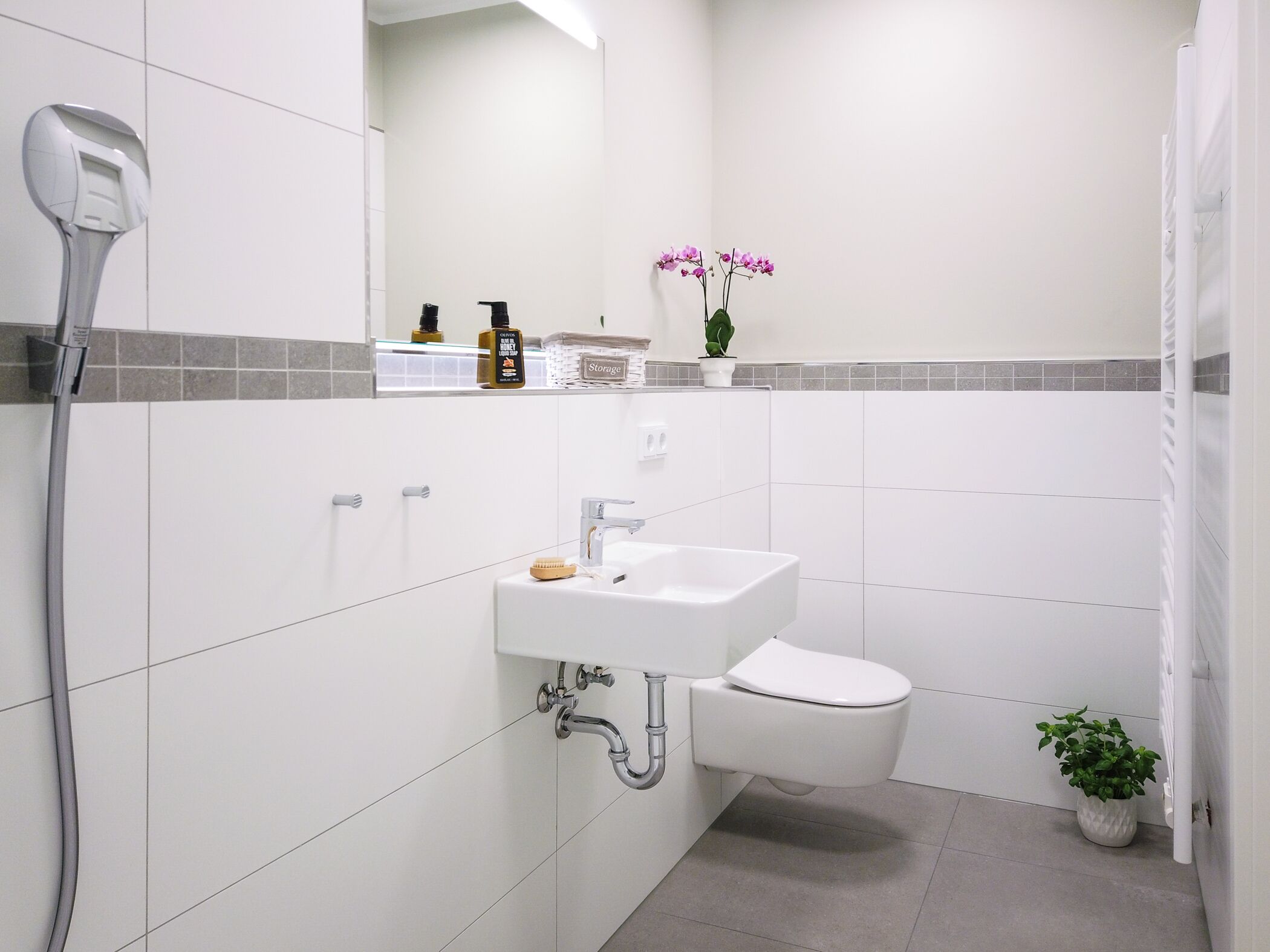 Ein modernes Badezimmer mit einem weißen Waschbecken, einer Toilette und einer Dusche. Über dem Waschbecken befindet sich ein Spiegel und in der Nähe der Toilette ein kleiner Topfpflanze.