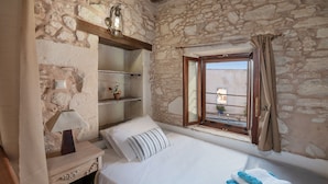 A bedroom with stone walls, a wooden nightstand, a bedside table with a lamp, a bed with white bedding, a window with curtains, and a view of the outside. - Kritiko Arhontiko (Apokoronas)