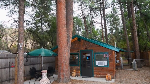 A rustic cabin with a green roof, a door with an American flag, and a small porch area with a table and chairs. - River Cabin (Ruidoso)