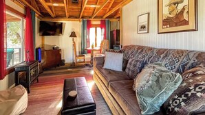 Un salon chaleureux avec un canapé en cuir, des poutres apparentes en bois au plafond, un téléviseur et des œuvres d’art encadrées accrochées aux murs.