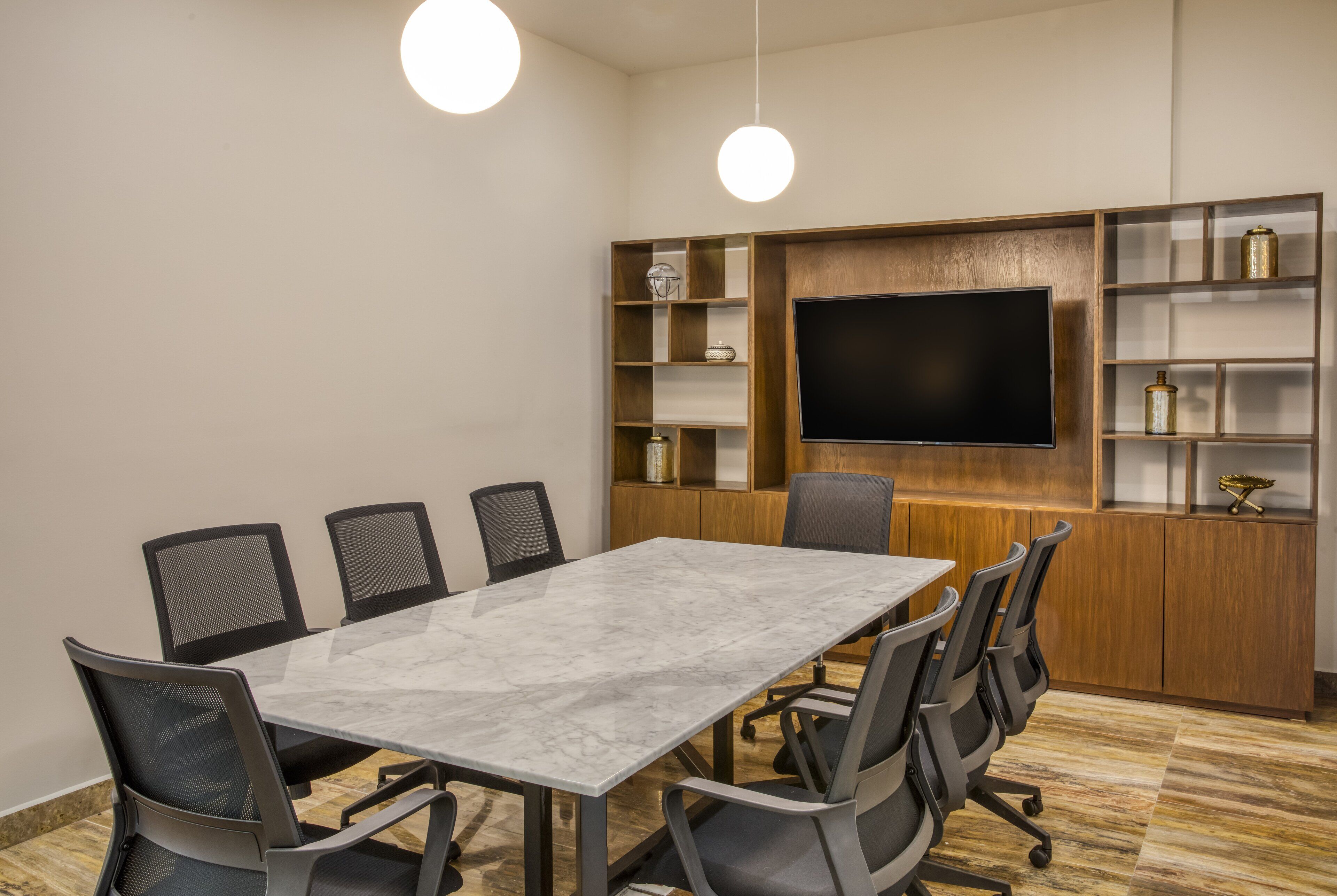 A conference room with a marble table, black chairs, a large flat-screen TV, wooden shelving with decorative items, and pendant lighting.