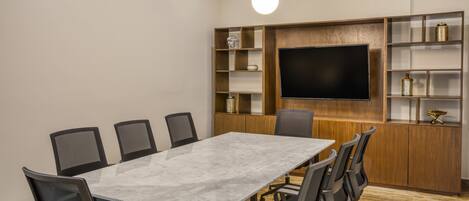 A conference room with a marble table, black chairs, a large flat-screen TV, wooden shelving with decorative items, and pendant lighting.