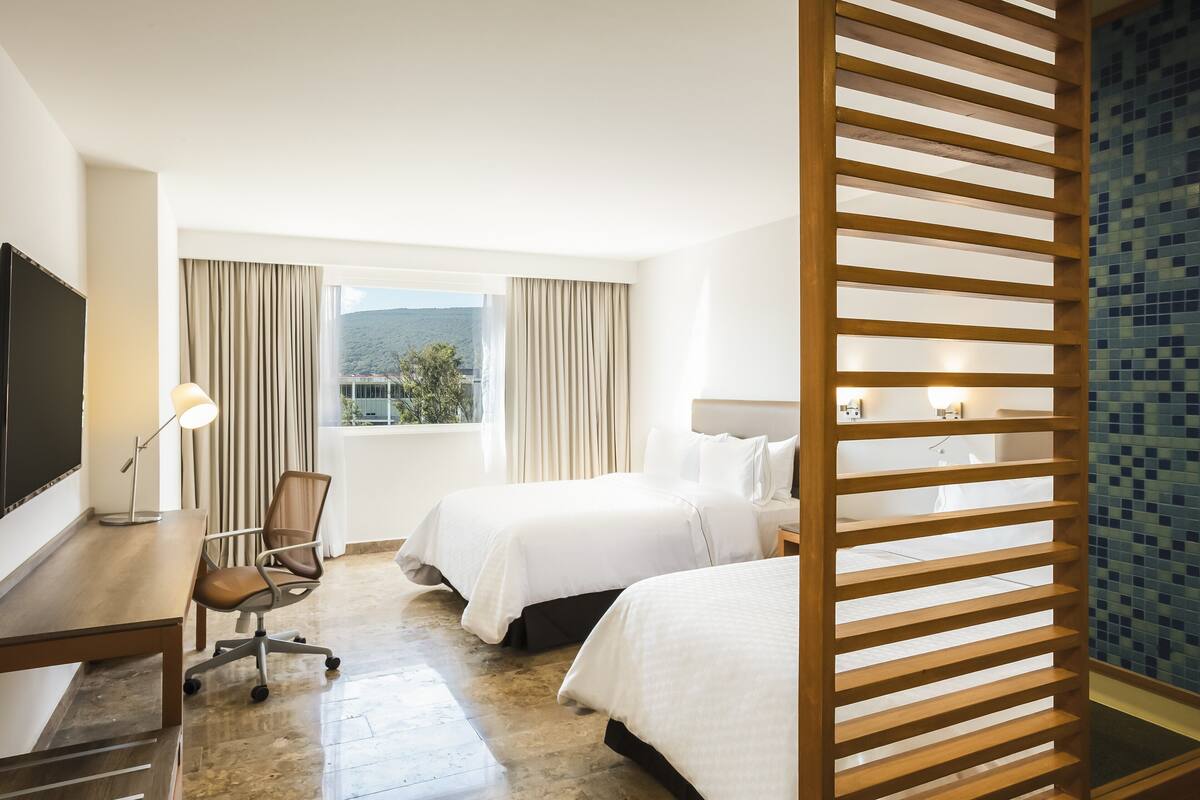 A hotel room with two beds, a desk with a chair, a TV, and a window with curtains.