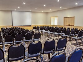 Un salón de conferencias con filas de sillas, una pantalla para proyector y una puerta de madera.