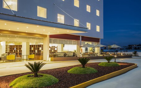 A modern hotel exterior with a well-lit entrance, outdoor seating area, and landscaped garden beds.