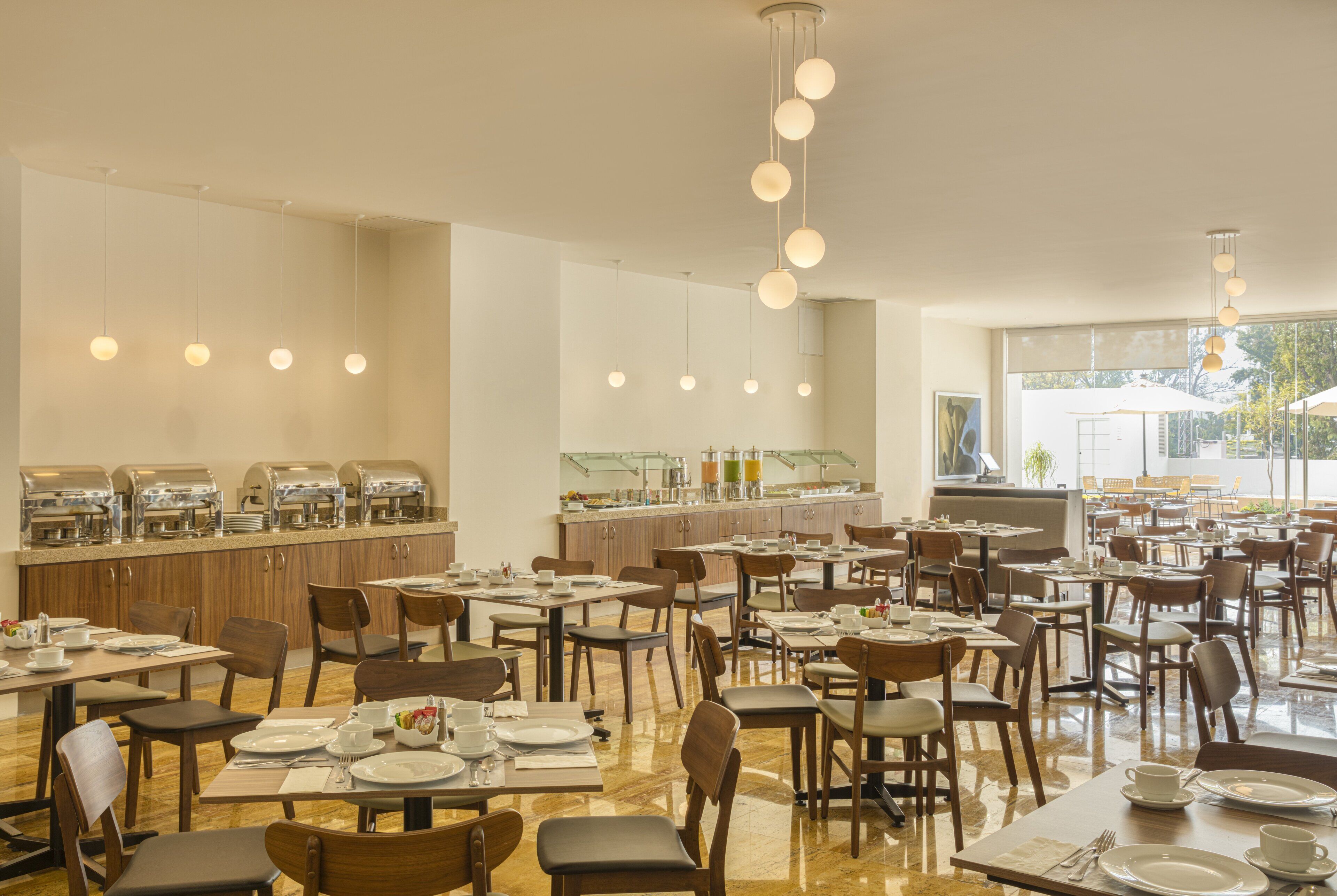 Un comedor amplio con varias mesas y sillas, un mostrador de buffet con diferentes alimentos, y lámparas colgantes.