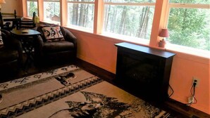 Ein gemütliches Wohnzimmer mit Kamin, großen Fenstern mit Blick auf einen Wald, einem gemusterten Teppich und einer bequemen Couch.