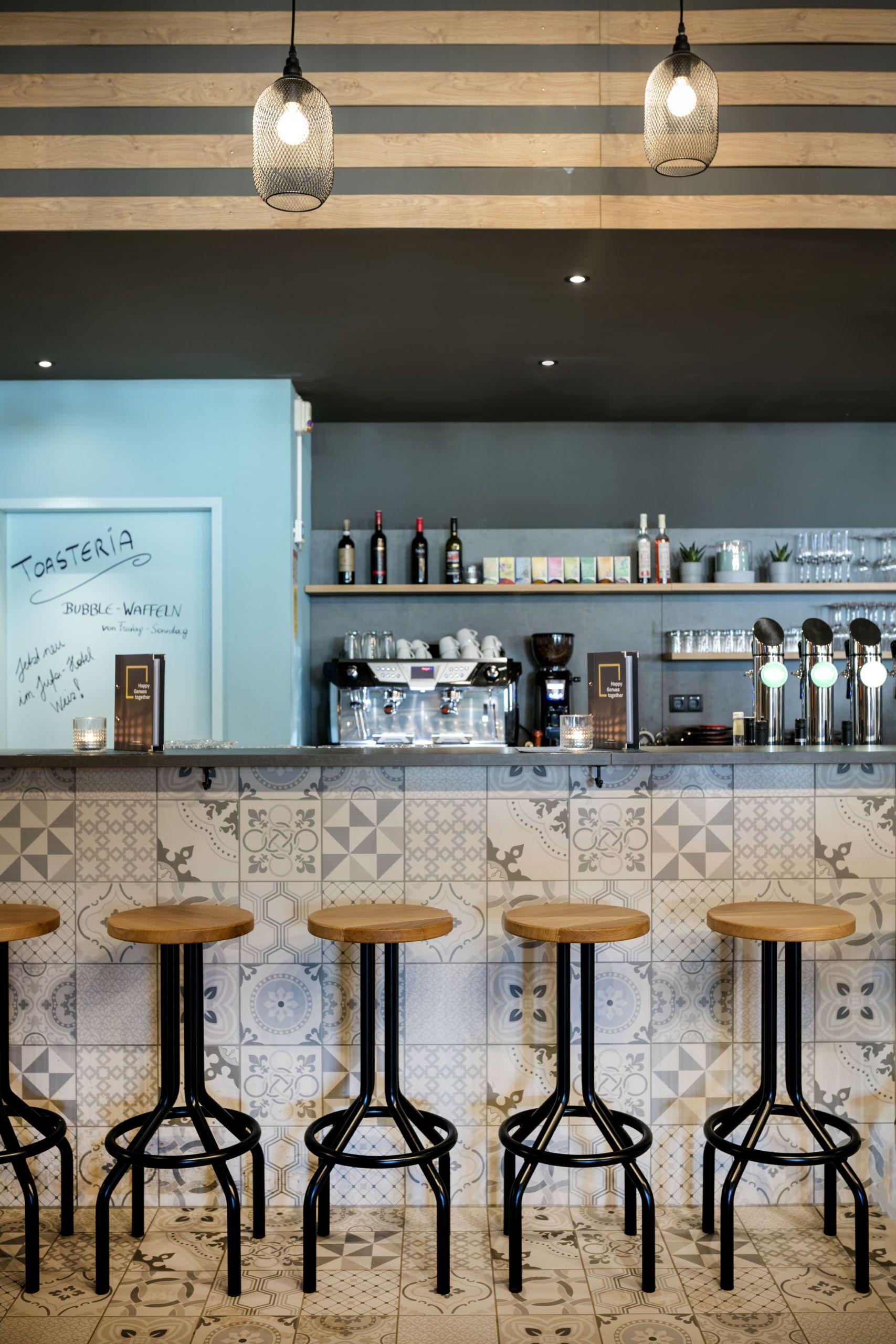 Un espace bar avec un mur recouvert de carreaux, des tabourets en bois, des suspensions décoratives et une machine à café.