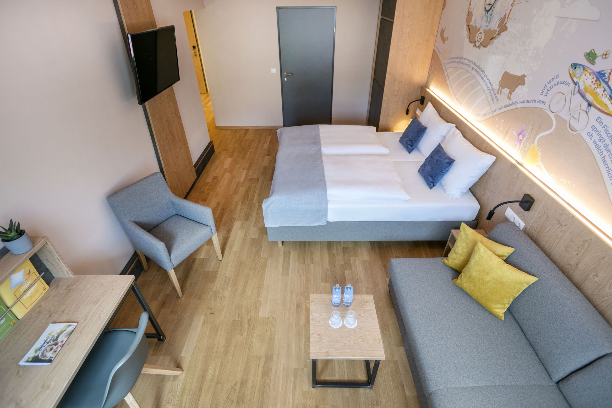 A modern hotel room with a sofa bed, a small table with water bottles, a chair, and a TV mounted on the wall.