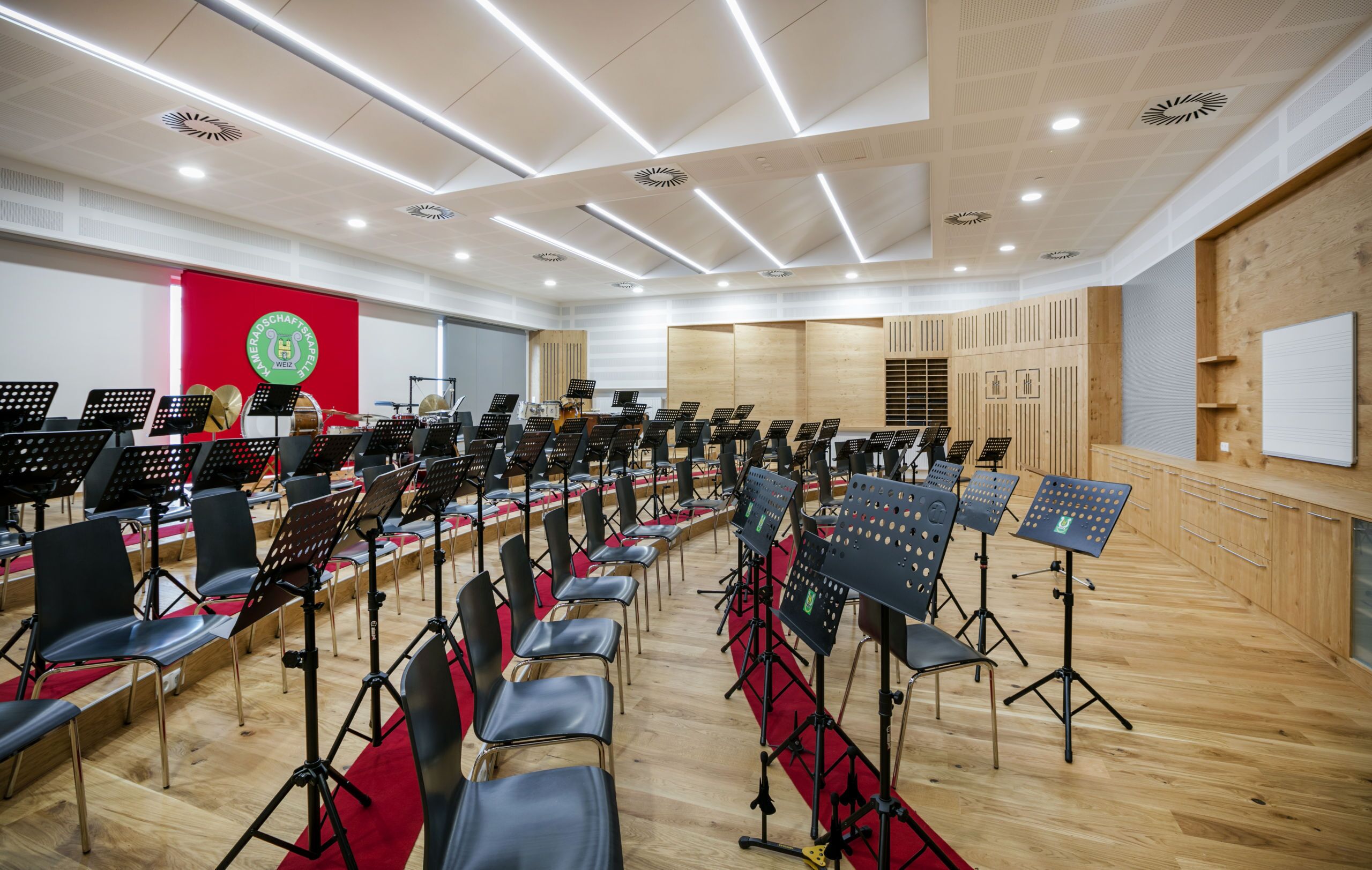 Une grande salle de concert, avec des rangées de chaises et des pupitres, un mur rouge orné d’un logo et des lambris en bois sur les murs.
