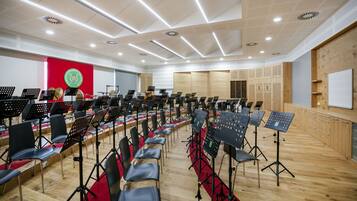 Un amplio salón de música con hileras de sillas y atril, una pared roja con un logotipo y paneles de madera en las paredes.