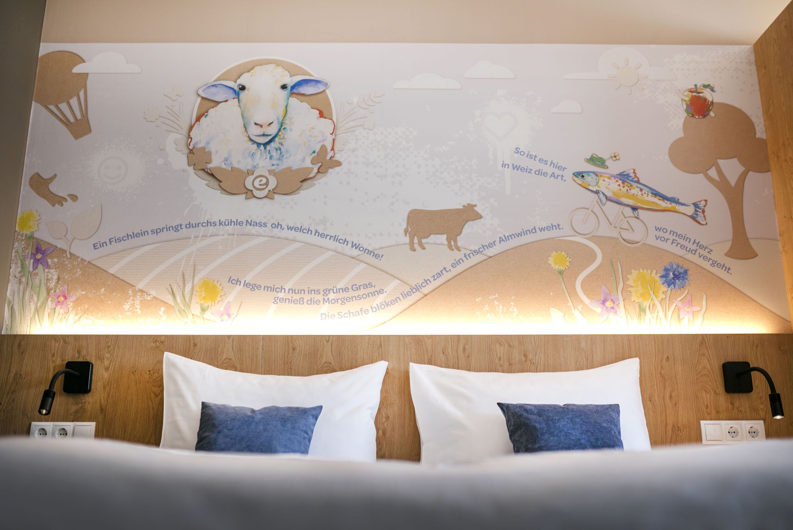Habitación de hotel con cabecera de madera, dos almohadas blancas y un mural que representa una oveja, un pez, una vaca y una bicicleta.