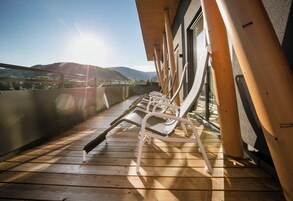 Um terraço com soalho de madeira banhado pelo sol e cadeiras de metal brancas, com vista para uma paisagem montanhosa.