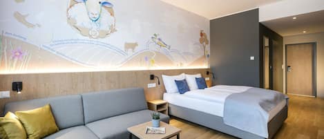 A modern hotel room with a grey sofa, a bed with a grey duvet, a small wooden table, and a wall mural depicting farm animals and a tractor.