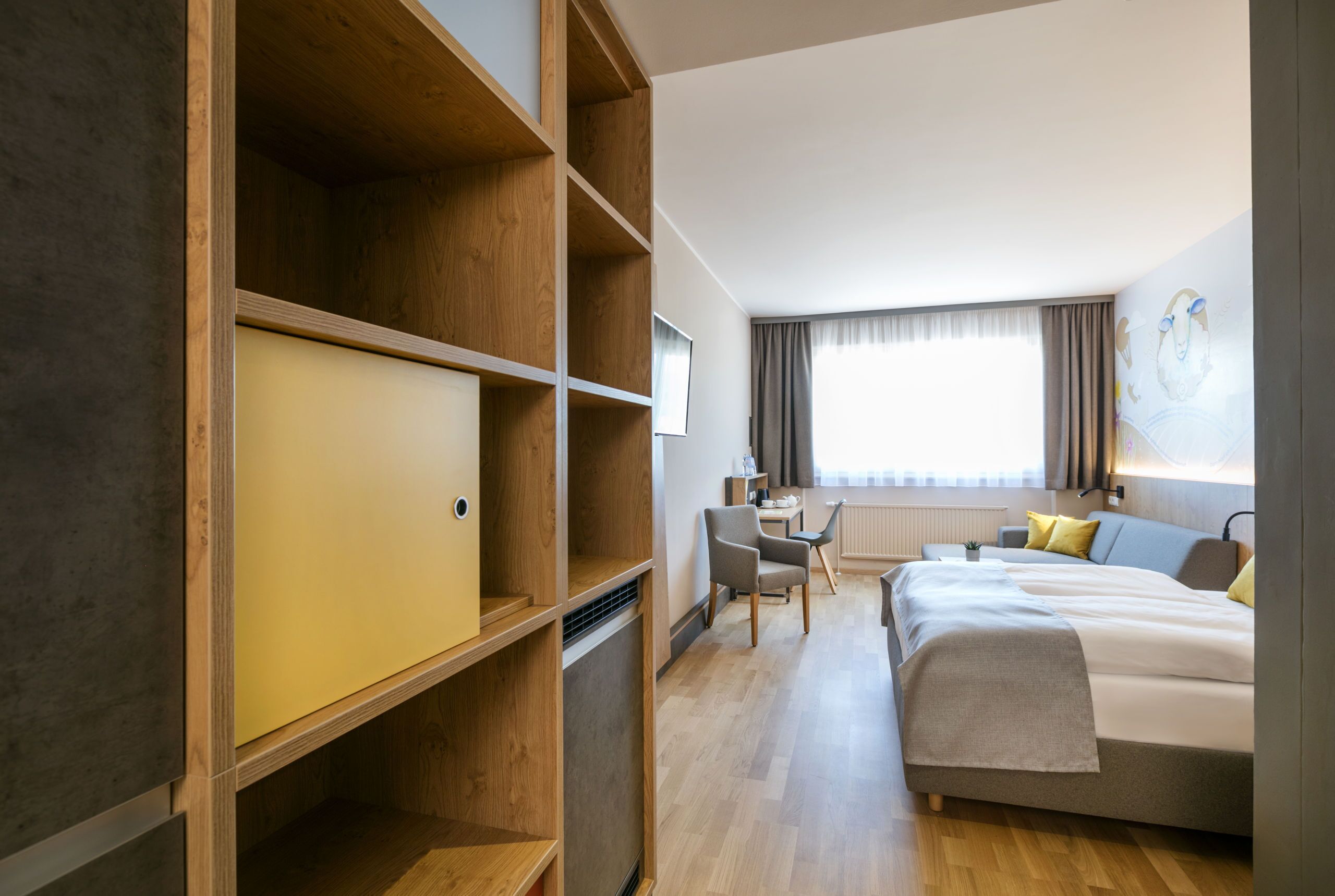 A hotel room with a bed, a desk, and a chair. There is a large wardrobe and a window with curtains.