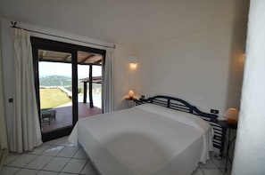 A bedroom with a bed, a nightstand, a lamp, and a view of a pool and landscape through a sliding door. - Villa Lara 2 (Arzachena)