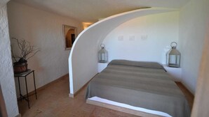 A bedroom with a curved ceiling, a bed, two wall-mounted lanterns, a small table with a potted plant, and a mirror on the wall. - Villa Lara 3 (Arzachena)