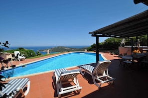 A swimming pool with lounge chairs and a table, overlooking a scenic view of the sea and hills.