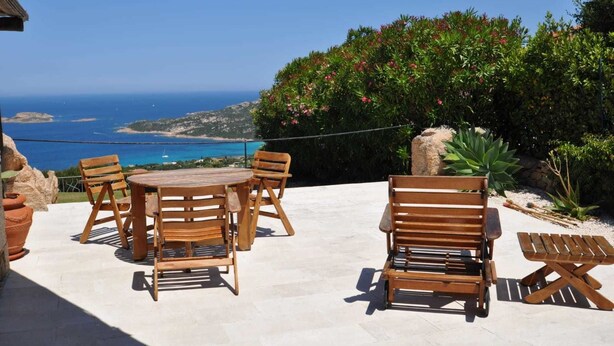 An outdoor patio with a round wooden table and chairs, a bench, and a view of the sea and hills. - Villa Lara 3 (Arzachena)