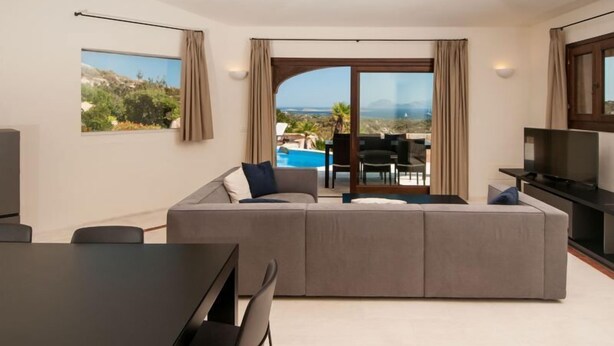 Ein modernes Wohnzimmer mit einer großen Couch, einem Esstisch und einem Fernseher. Durch die Glastüren ist ein Blick auf einen Pool und Berge möglich.
