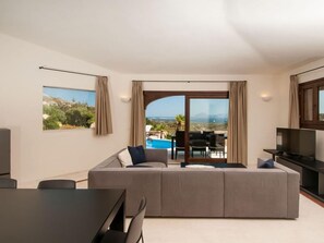 A modern living room with a large sofa, a dining table, and a TV. There is a view of a pool and mountains through the glass doors. - Villa Pevero Hills 1 (Arzachena)