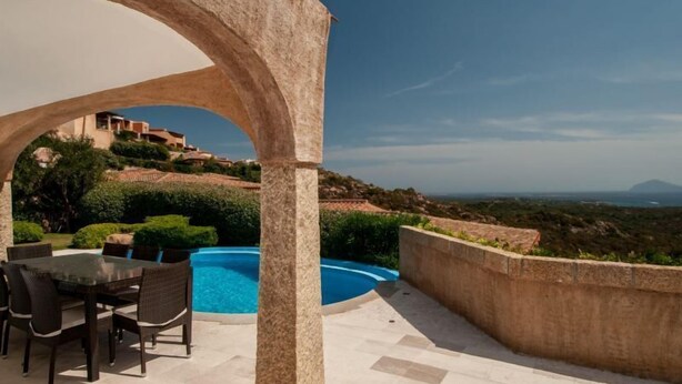 A pool area with a table and chairs, a stone archway, and a view of a hillside with buildings.