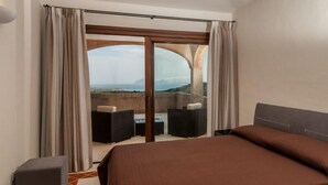 A hotel room with a large bed, a small sofa, a table with chairs, and a view of the sea through a glass door.