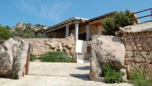 A stone entrance gate with the inscription "LU RISCIO" and a building with a tiled roof and columns. - Villa La Silvaredda (Arzachena)