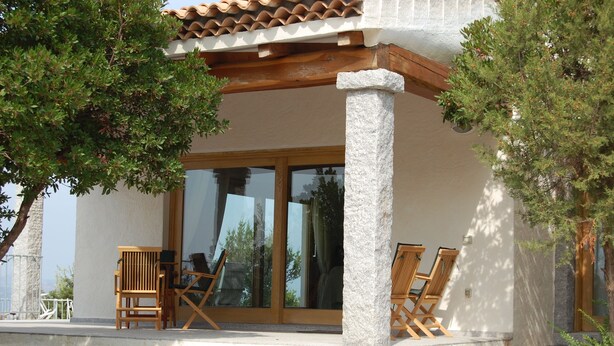 Un espace extérieur abrité, avec des chaises en bois et une table, un pilier en pierre et un bâtiment doté de grandes portes vitrées.
