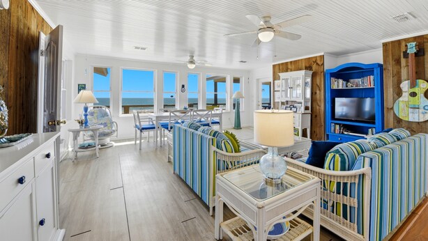 Ein geräumiges Wohnzimmer mit großem Fenster zum Meer, Deckenventilator und einer blauen Unterhaltungsanlage.