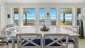Ein Zimmer direkt am Strand mit einem weißen Tisch und Stühlen, großen Fenstern mit Blick auf den Ozean und einem Deckenventilator.