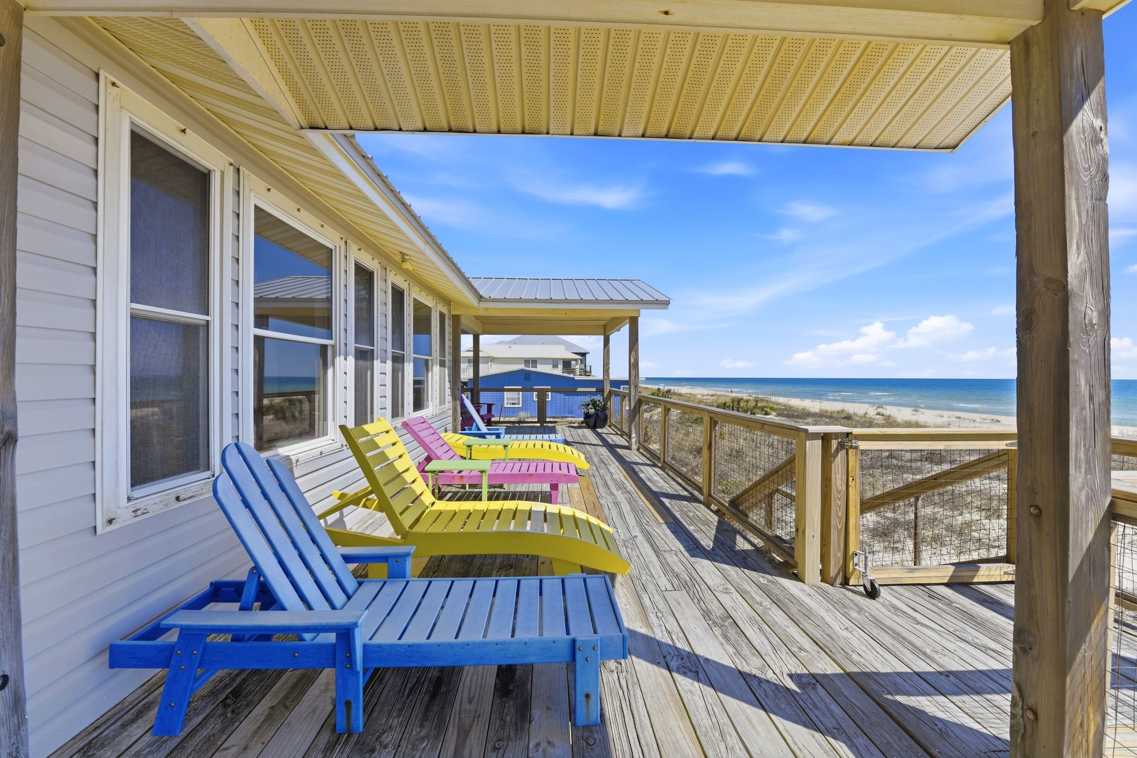 A wooden deck with blue and yellow Adirondack chairs facing a beach.