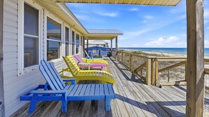 Eine Holzterrasse mit blau-gelben Adirondack-Stühlen, die auf einen Strand ausgerichtet sind.