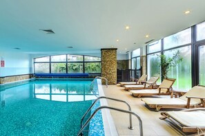 Una piscina interior con agua cristalina, rodeada de sillones de descanso y grandes ventanales con vistas a la vegetación.