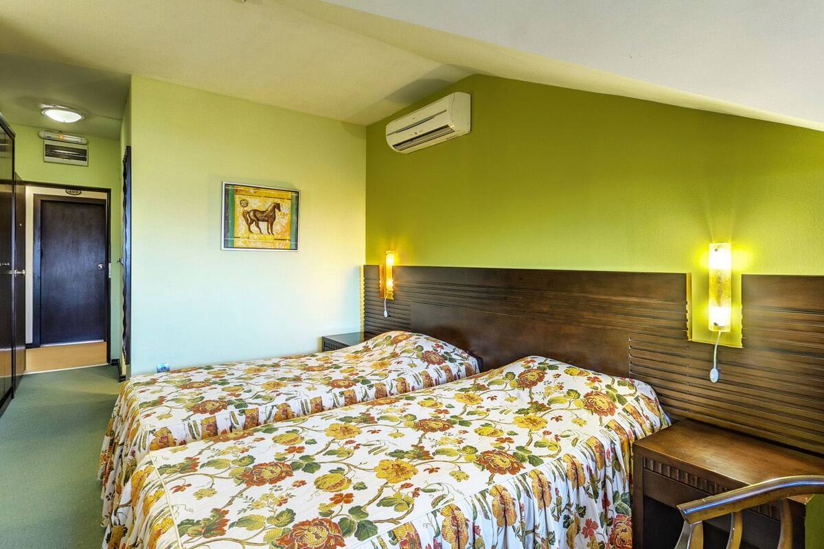 A hotel room with two beds, a wooden headboard, a wall-mounted air conditioner, a framed picture, and a desk with a chair.