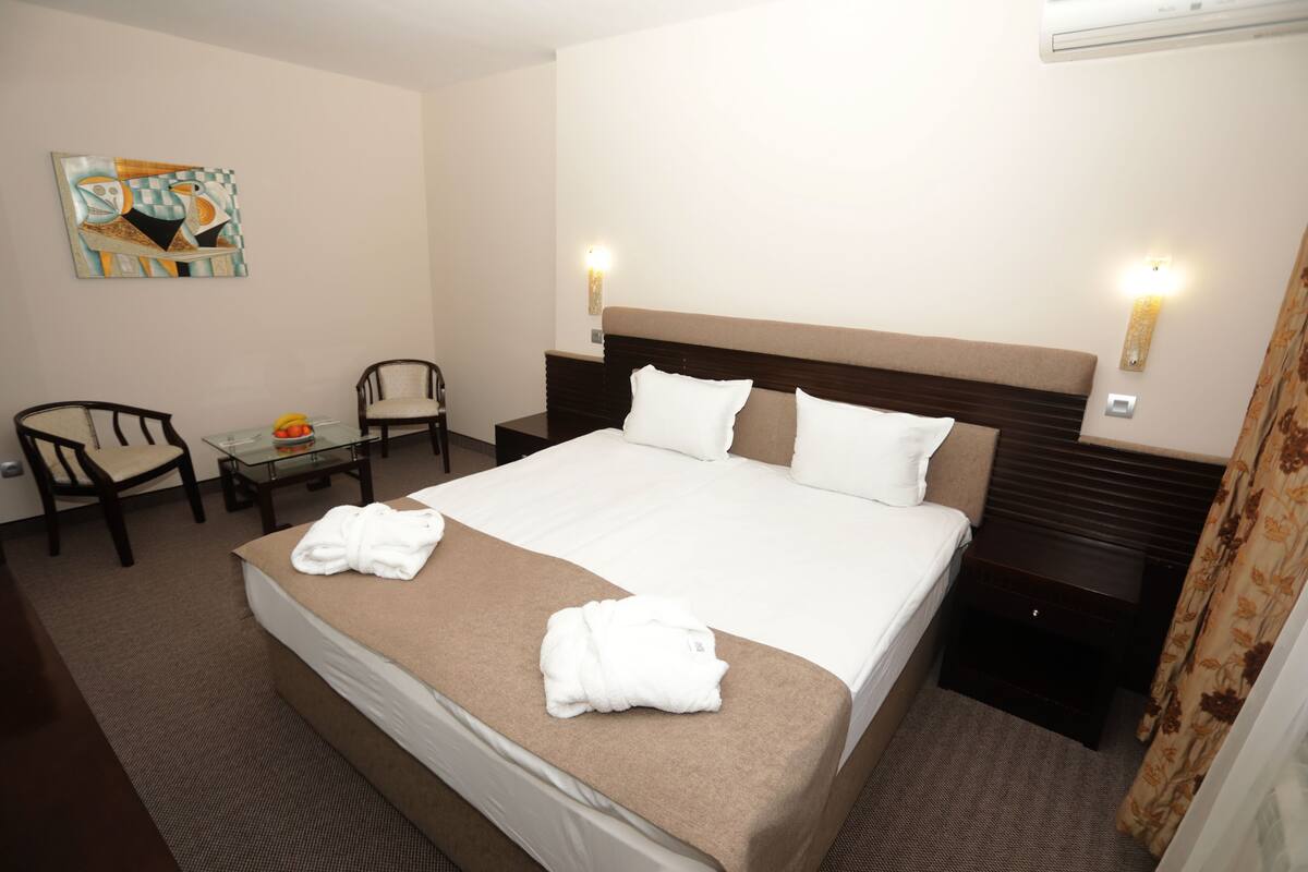 A hotel room with a large bed, two bedside tables, a chair, a small table with a fruit bowl, and a painting on the wall.