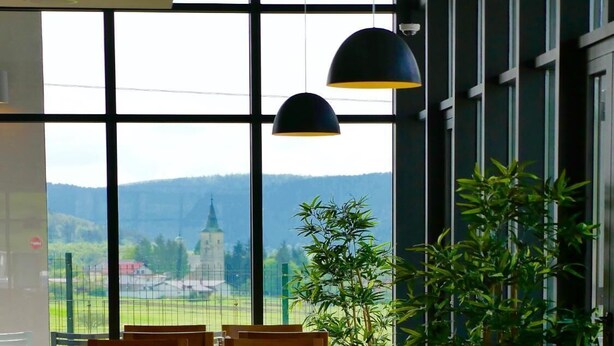 A restaurant with wooden chairs and tables, large windows offering a view of a distant town and church, and hanging pendant lights. - Hotel Lika Sjever (Brinje)