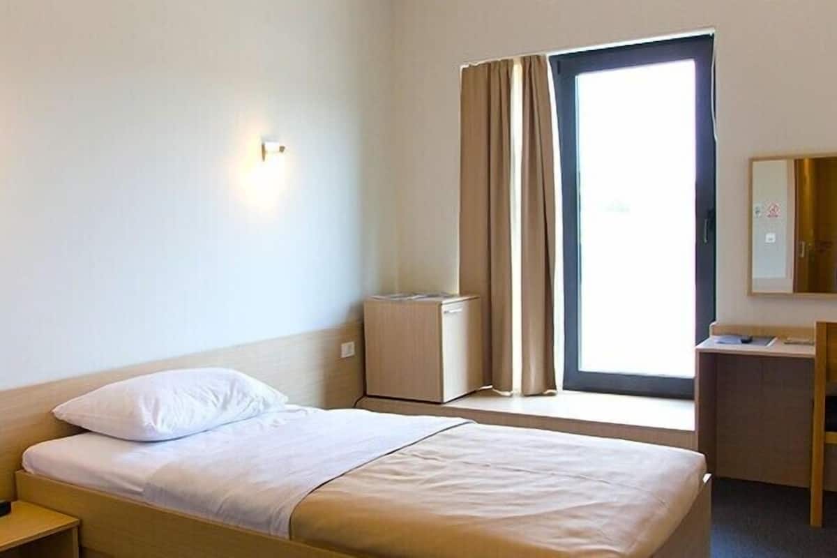 A hotel room with a bed, bedside table, lamp, wardrobe, desk, chair, and a window with curtains.