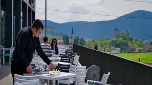 An outdoor dining area with white tables and chairs, overlooking a scenic landscape with hills and a church. - Hotel Lika Sjever (Brinje)
