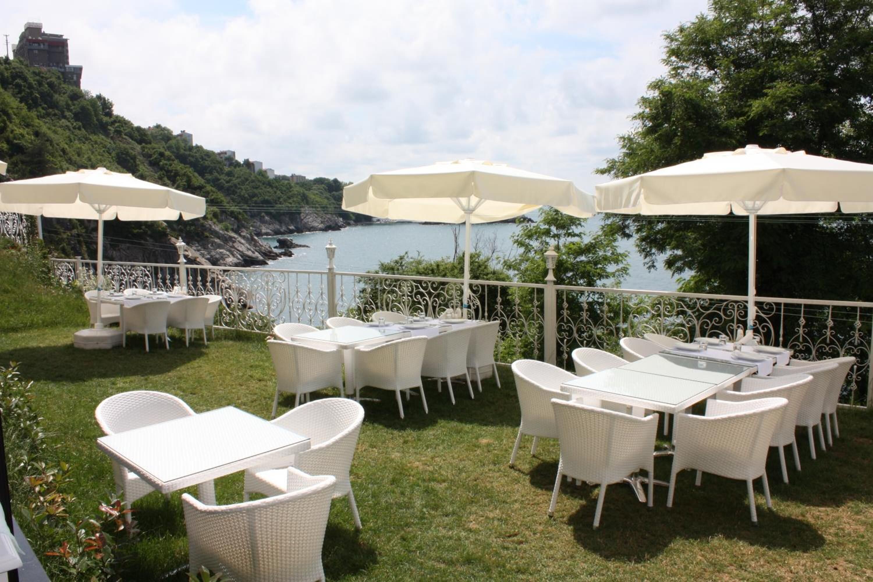 Une terrasse extérieure dotée de meubles en osier blanc et de parasols, offrant une vue sur une étendue d’eau et une côte rocheuse.