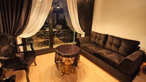 Un salon avec un canapé noir, une table ronde surmontée d’un plateau en marbre et un fauteuil. Des rideaux sont accrochés aux fenêtres, et on aperçoit la rambarde d’un balcon.