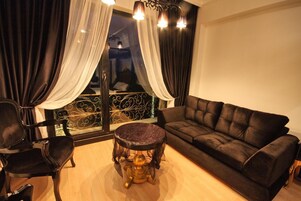 A living room with a black sofa, a round table with a marble top, and a chair. There are curtains on the windows, and a balcony railing is visible.