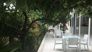Un patio al aire libre con mobiliario de mimbre blanco y suelo de baldosas, rodeado de abundante vegetación y un edificio blanco al fondo.