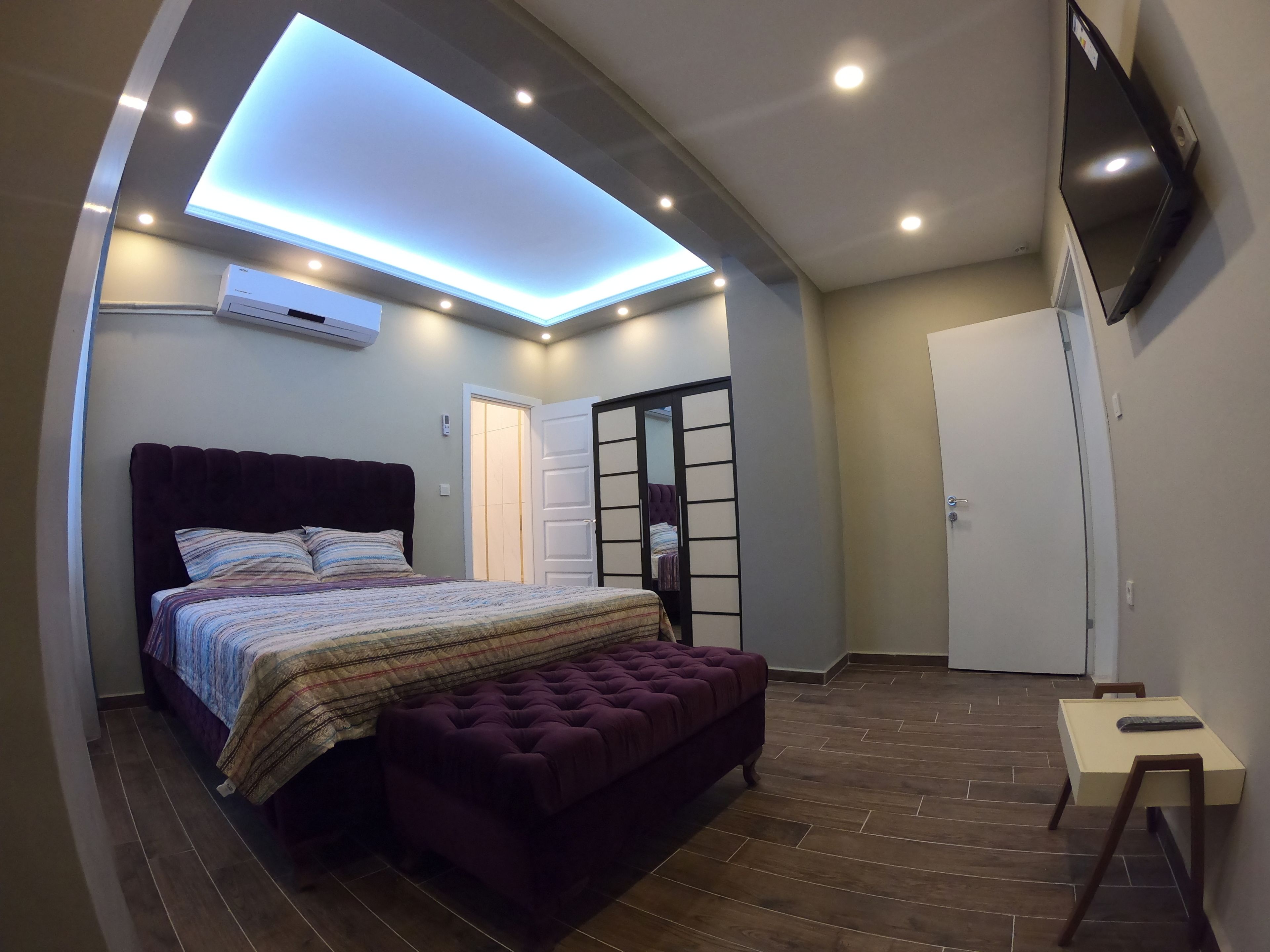 A bedroom with a bed, a tufted bench, a small table, a TV, and a ceiling light fixture.