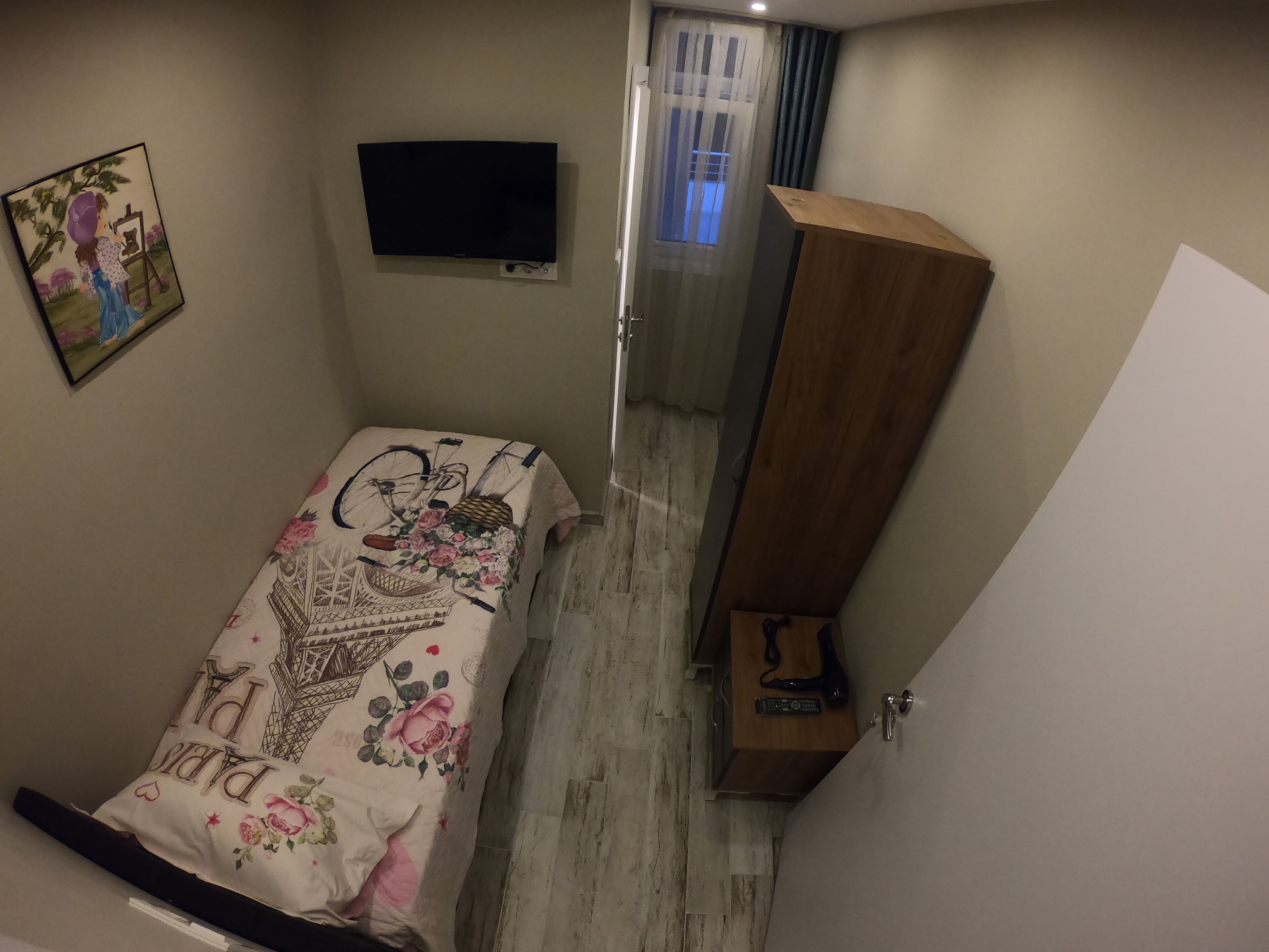 Une petite chambre à coucher comprenant un lit, un téléviseur fixé au mur, une armoire en bois et une porte donnant sur un balcon.