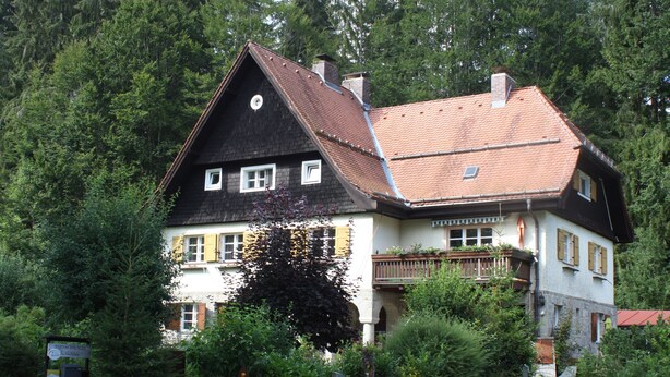 Une maison à deux étages, dotée d’un toit à forte pente, entourée d’une forêt dense.