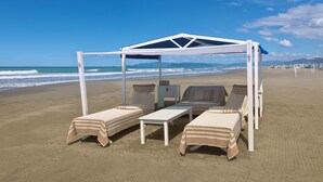 A beach cabana with two lounge chairs and a table, set on a sandy beach with the ocean in the background. - Augustus Lido (Forte dei Marmi)