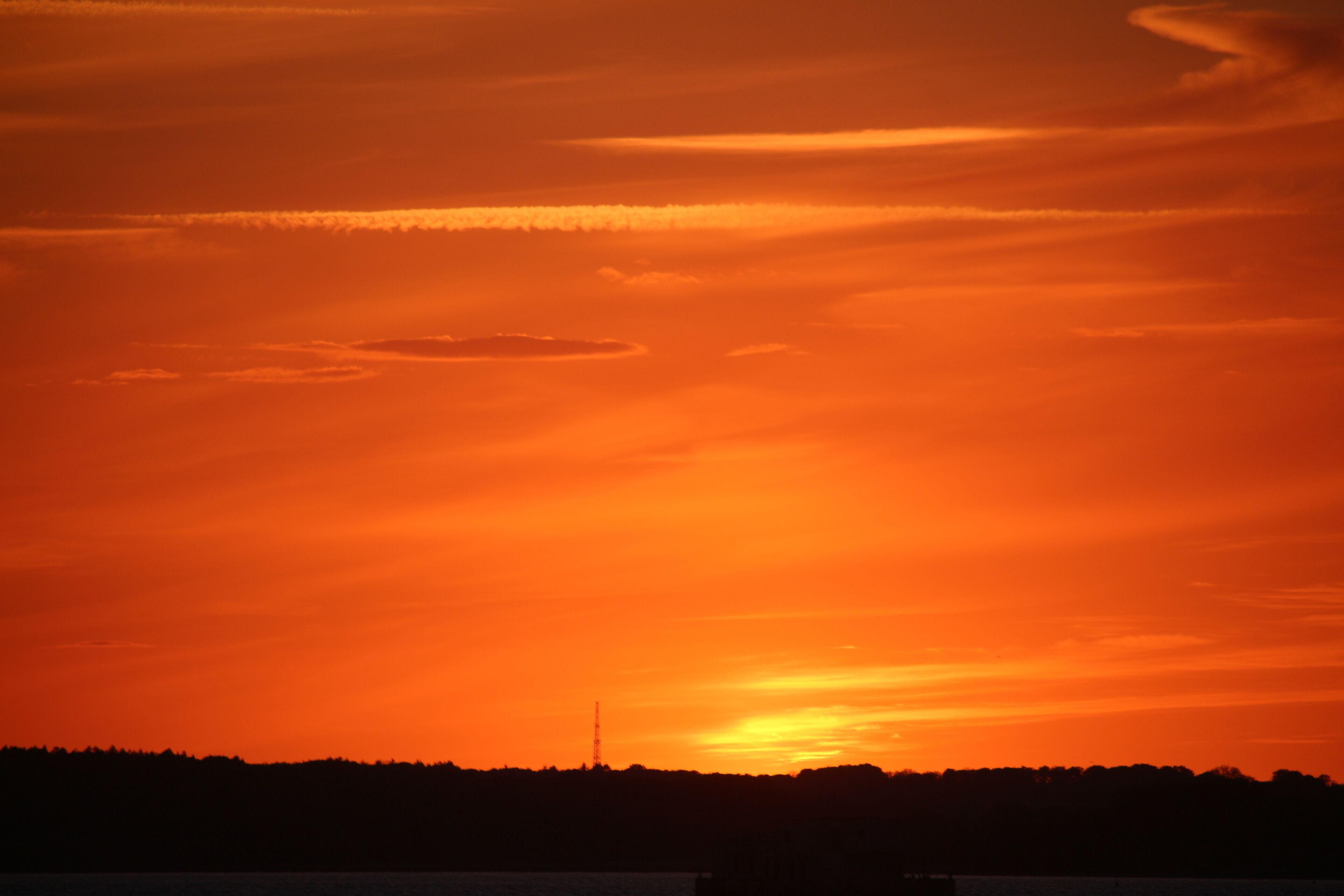 A sunset over a body of water with a silhouette of a forested area and a distant tower.