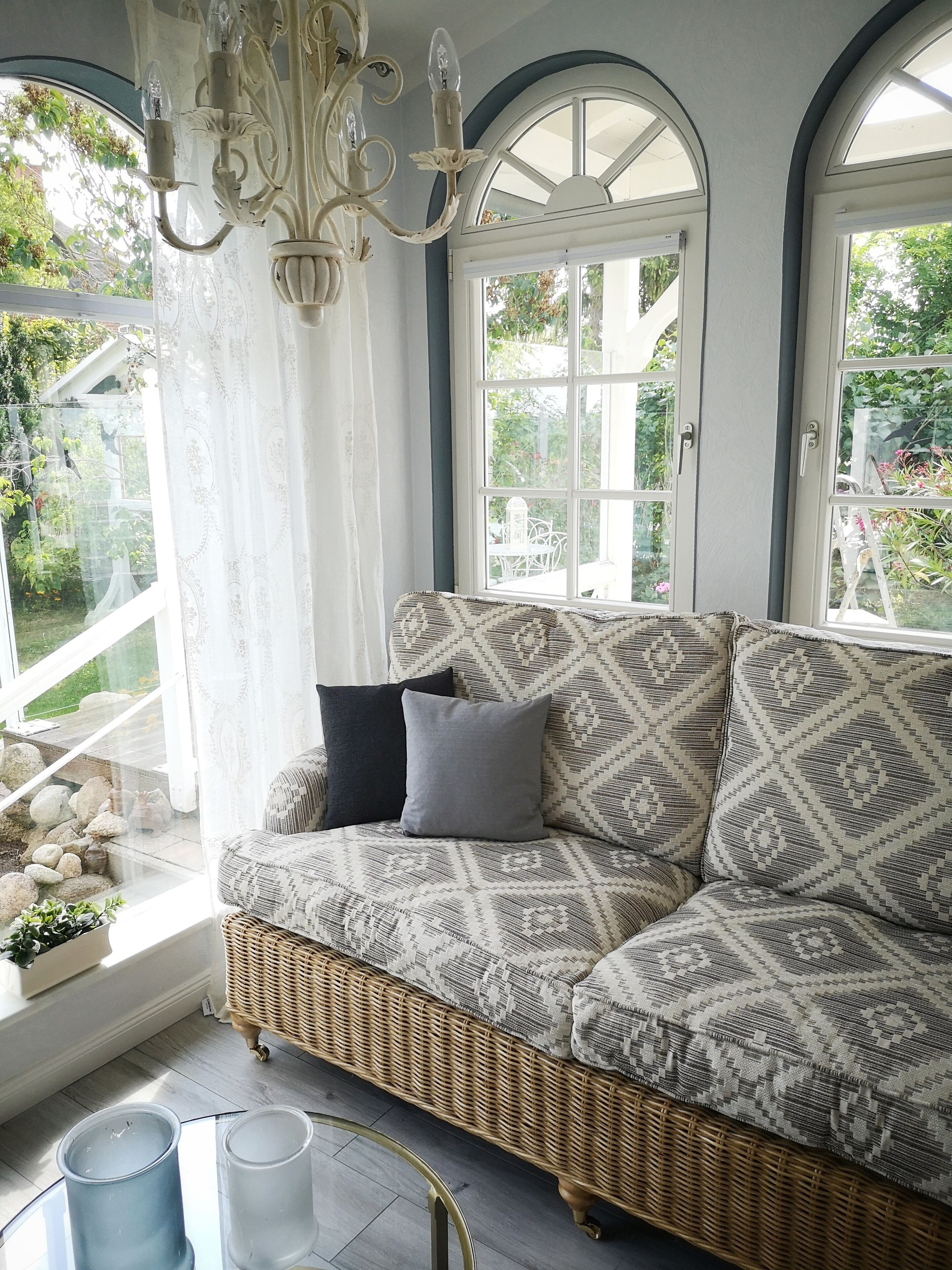 A wicker sofa with patterned cushions, a chandelier, and a view of greenery through large windows.