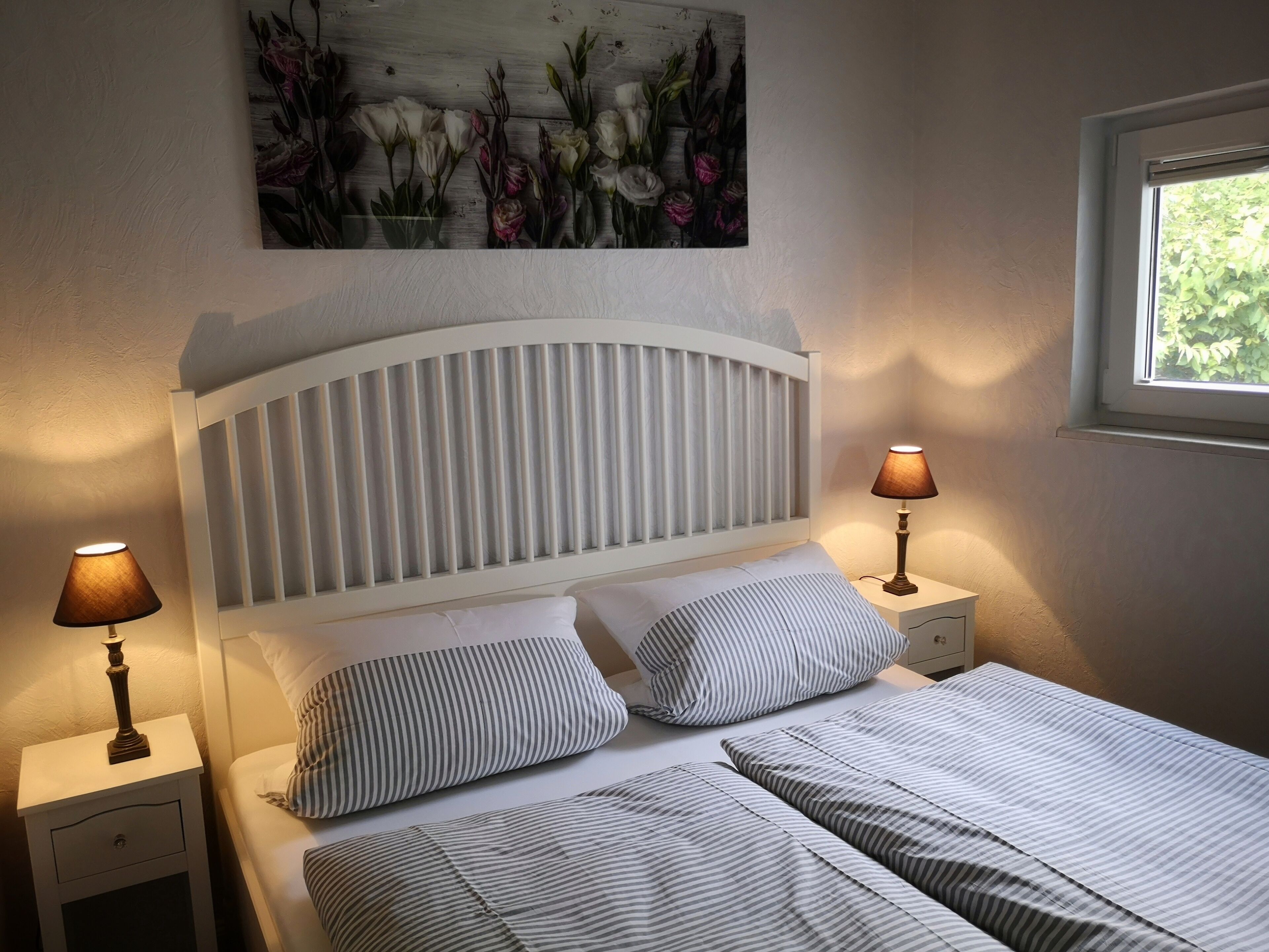 A bedroom with a white headboard, two bedside lamps, striped bedding, and a floral painting on the wall.